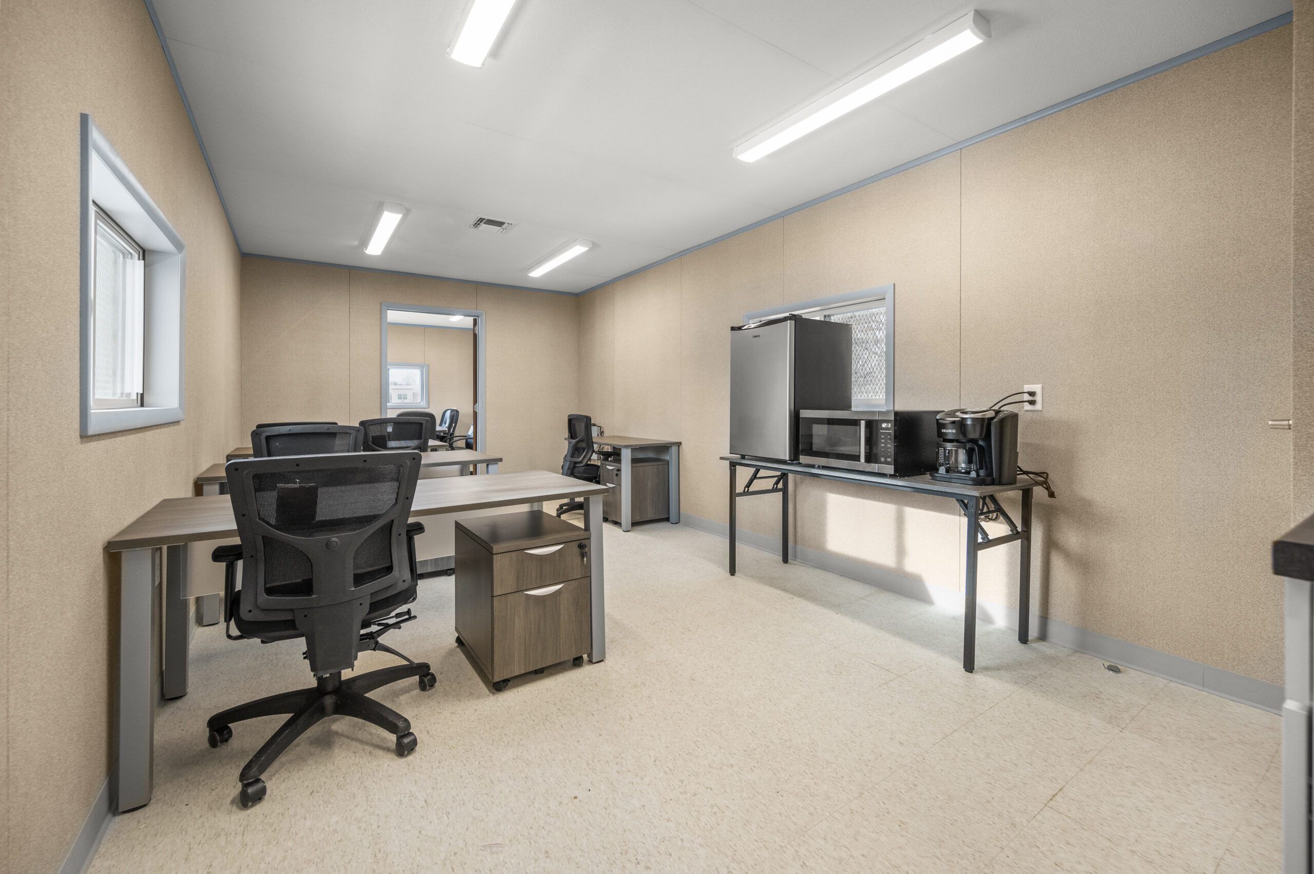 Interior of an office with desks, chairs, and a kitchenette area featuring a refrigerator and microwave.