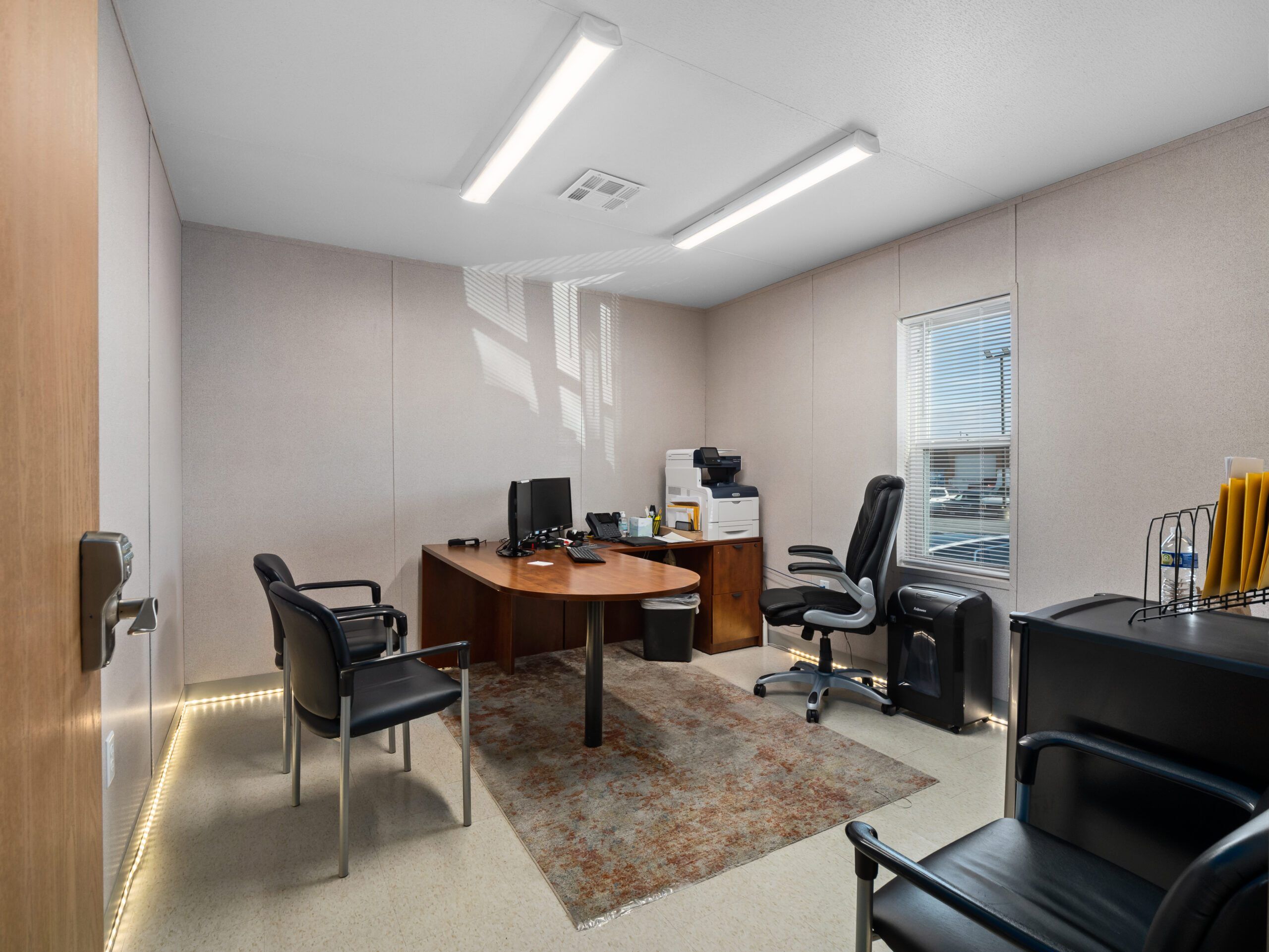 A modern office interior with a wooden desk, black chairs, and a printer, lit by fluorescent lights and natural light from a small window.