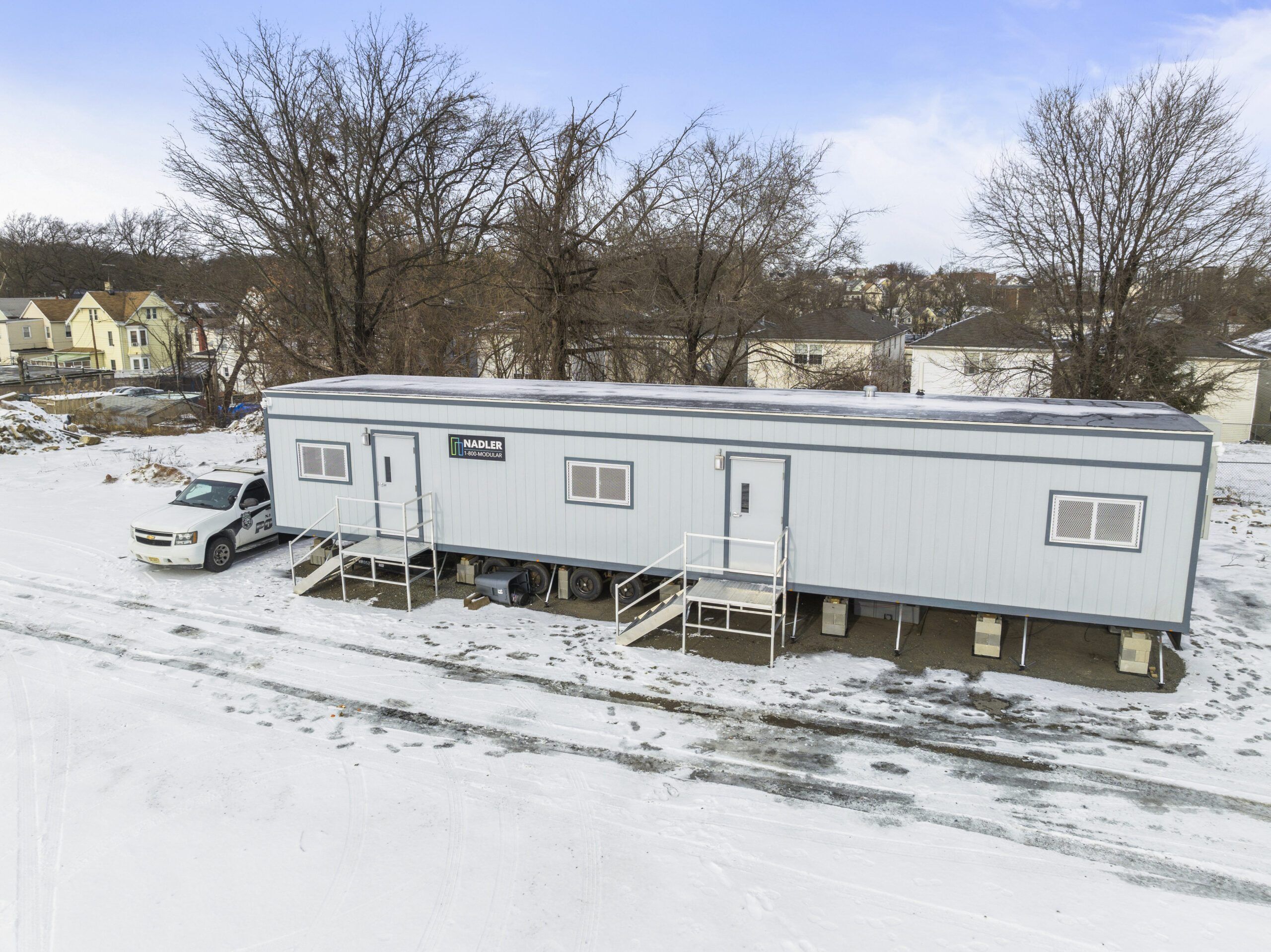 Modular office trailer set up on a snowy lot, with Nadler Modular branding visible on the side. A vehicle is parked nearby, and residential homes are visible in the background.